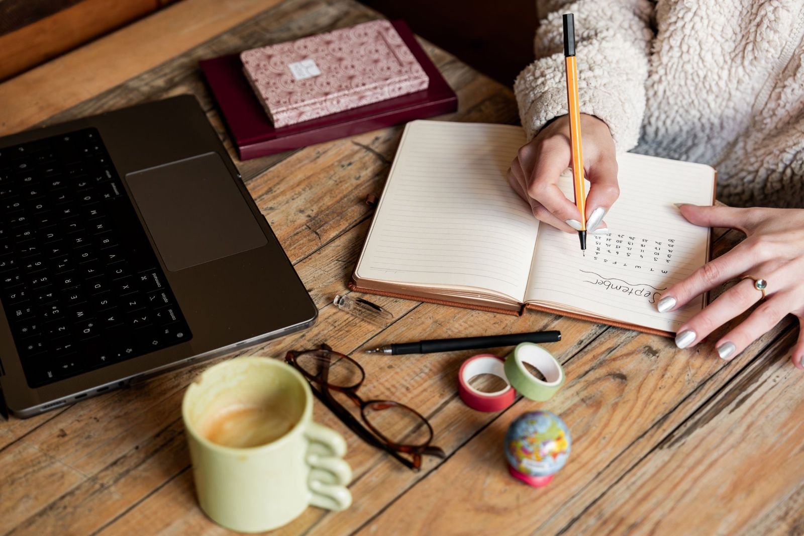 Hands writing a monthly plan in a notebook beside a laptop, coffee mug and glasses on a wooden table
